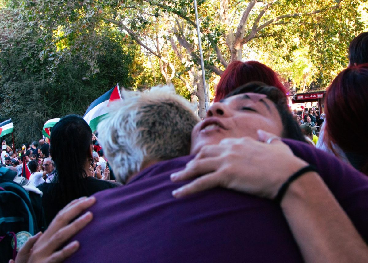 Two protestors embrace in solidarity while the main crowd roars from afar.