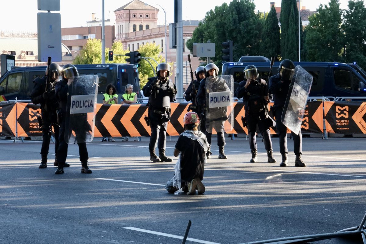 A pro-Palestine protester kneels in prayer in front of guarded police while officers scan the crowd for retaliation. 